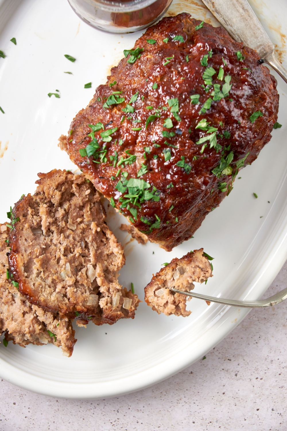 A sliced BBQ meatloaf garnished with parsley on a white serving tray. A fork picks up a piece of meatloaf.