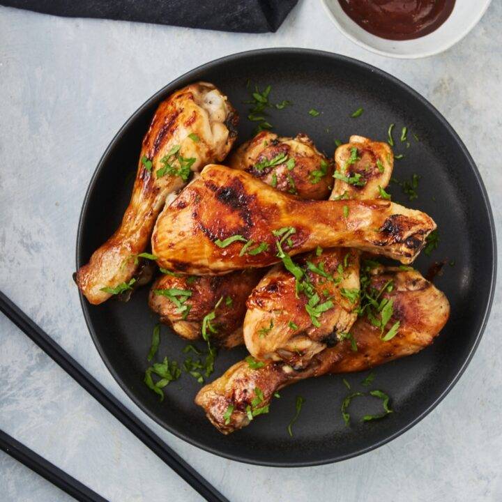 A pile of barbecue chicken drumsticks on a black plate garnished with fresh herbs and a bowl of barbecue sauce is next to it.