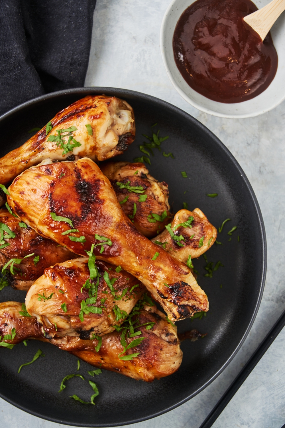 A pile of barbecue chicken drumsticks on a black plate garnished with fresh herbs and a bowl of barbecue sauce is next to it.