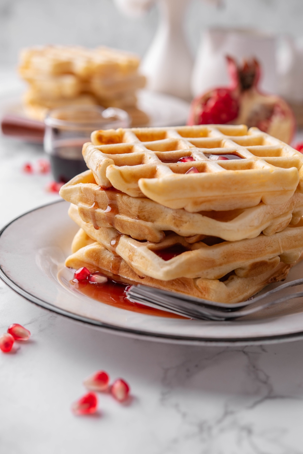 Four waffles stacked on top of each other with maple syrup dripping down the sides of the waffles and pomegranate seeds sprinkled on top and around the waffles. There are two forks on the plate.