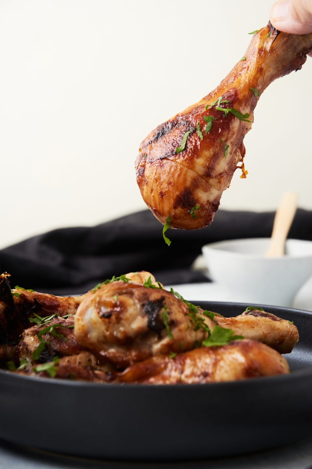 A barbecue chicken drumstick being removed from a plate filled with drumsticks. Bits of fresh herbs are garnished on the chicken.