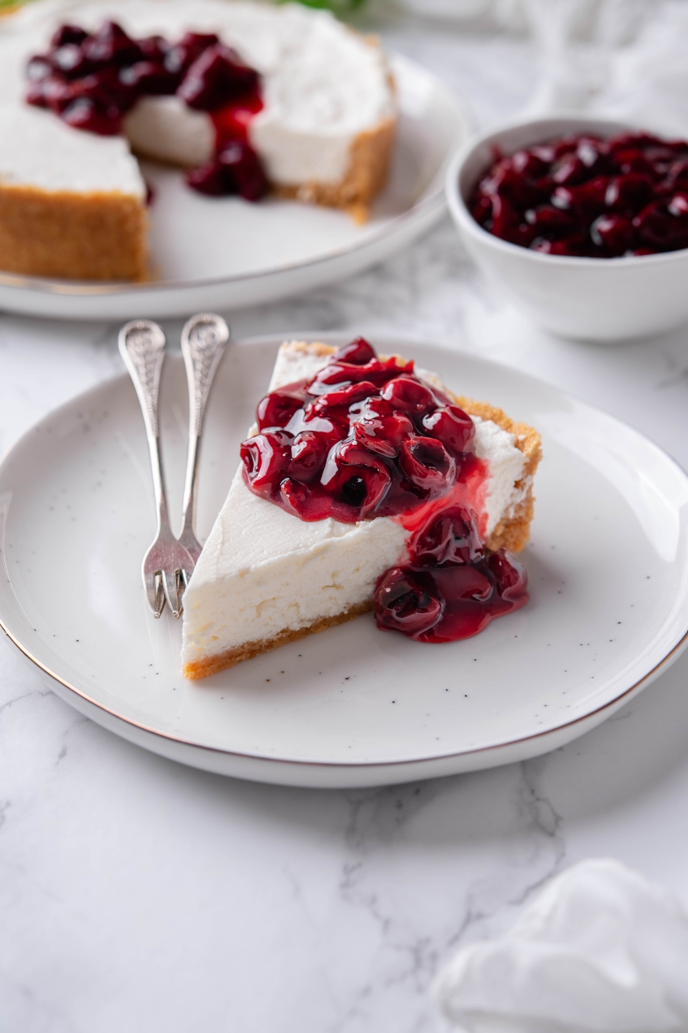 A serving of cream cheese pie topped with cherry pie filling on a white plate with two forks on the plate.