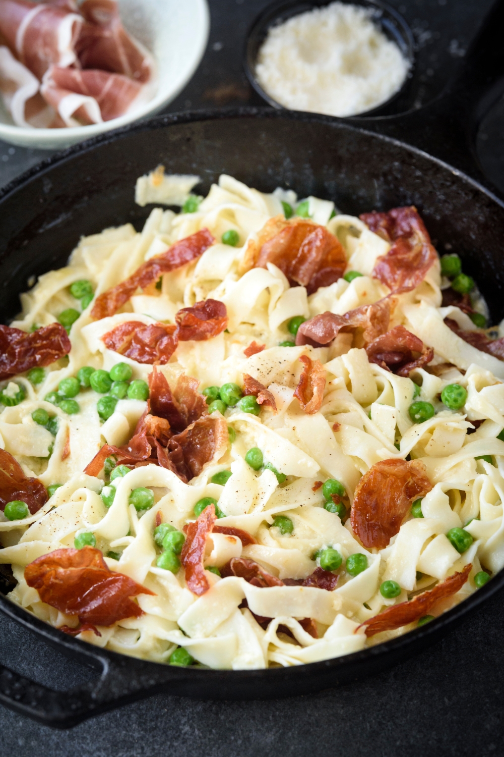 A cast iron skillet filled with cooked pasta mixed with peas and prosciutto in a creamy sauce.