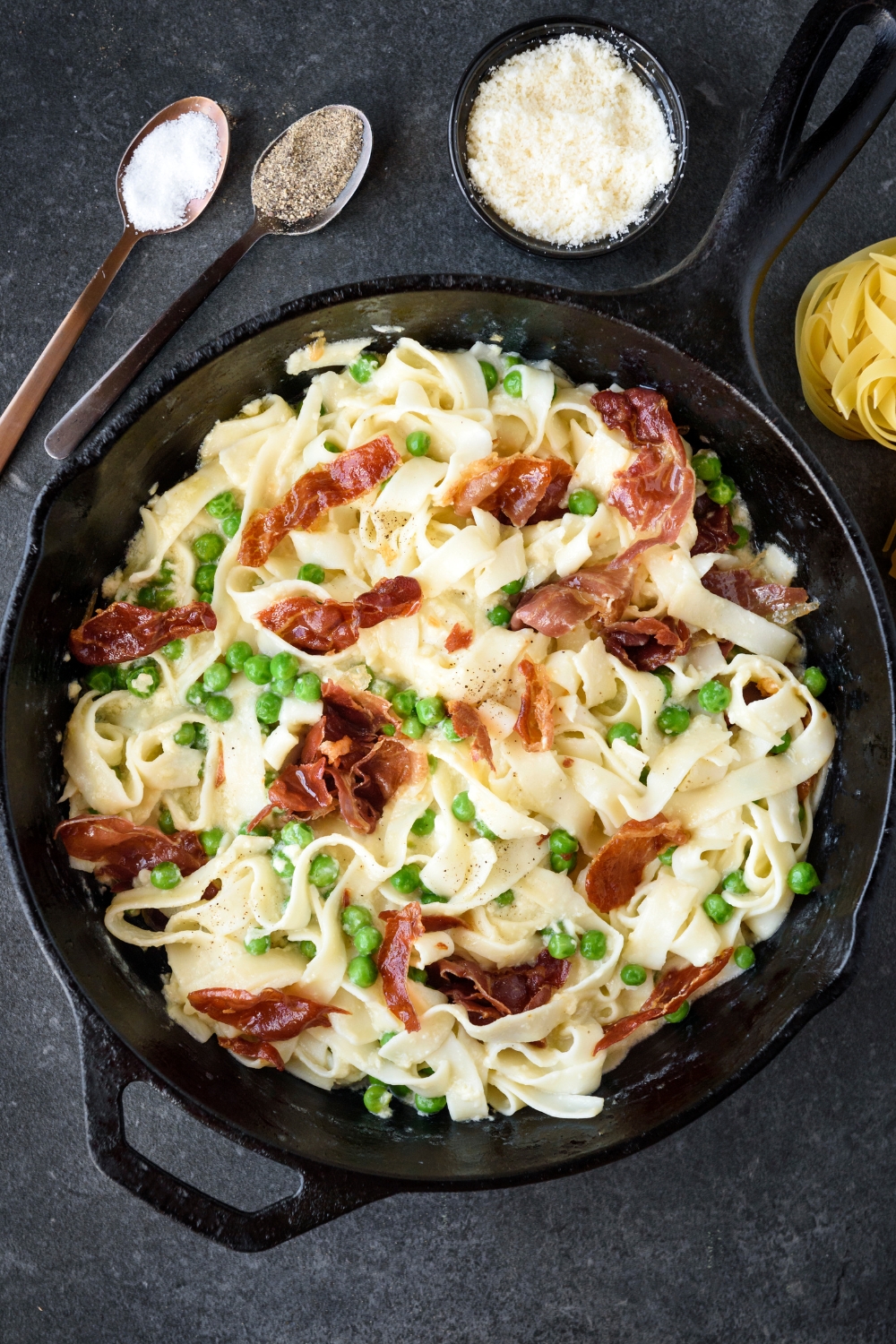 A cast iron skillet filled with cooked pasta mixed with peas and prosciutto in a creamy sauce.