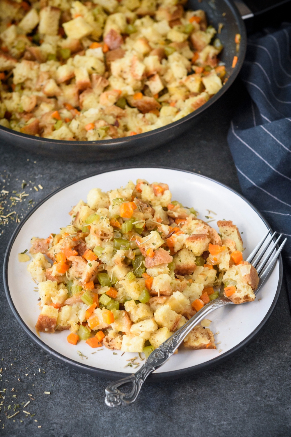 A plate of stovetop stuffing with diced celery and carrots mixed in with the stuffing. There is a fork on the plate and the rest of the stuffing is in a skillet in the background.