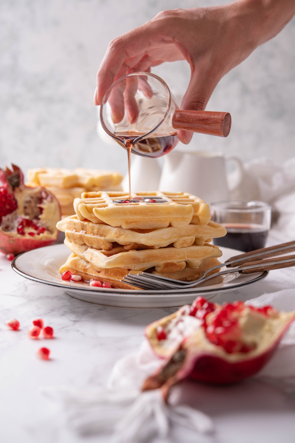 A hand pouring a jar of maple syrup over top of four waffles stacked on top of each other. There are two forks on the plate.