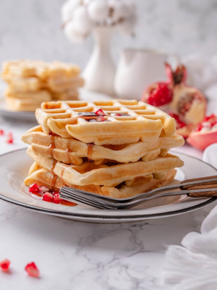 Four waffles stacked on top of each other with maple syrup dripping down the sides of the waffles and pomegranate seeds sprinkled on top and around the waffles.