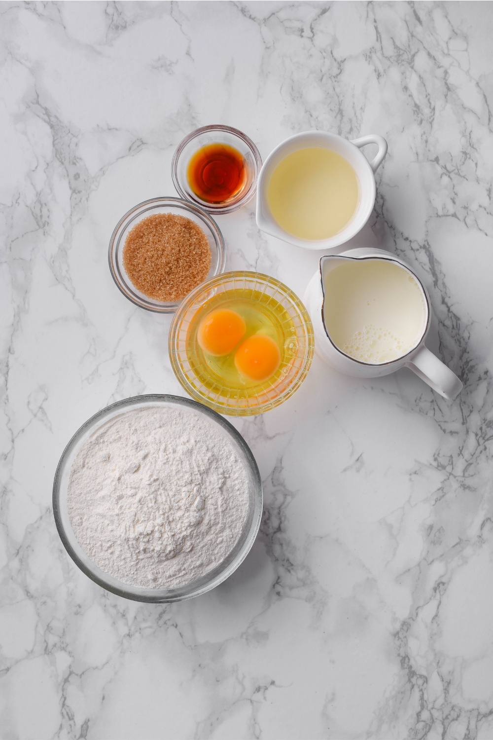 An assortment of ingredients including bowls of flour, unbeaten eggs, vanilla extract, brown sugar, milk, and oil, all on a white marble countertop.