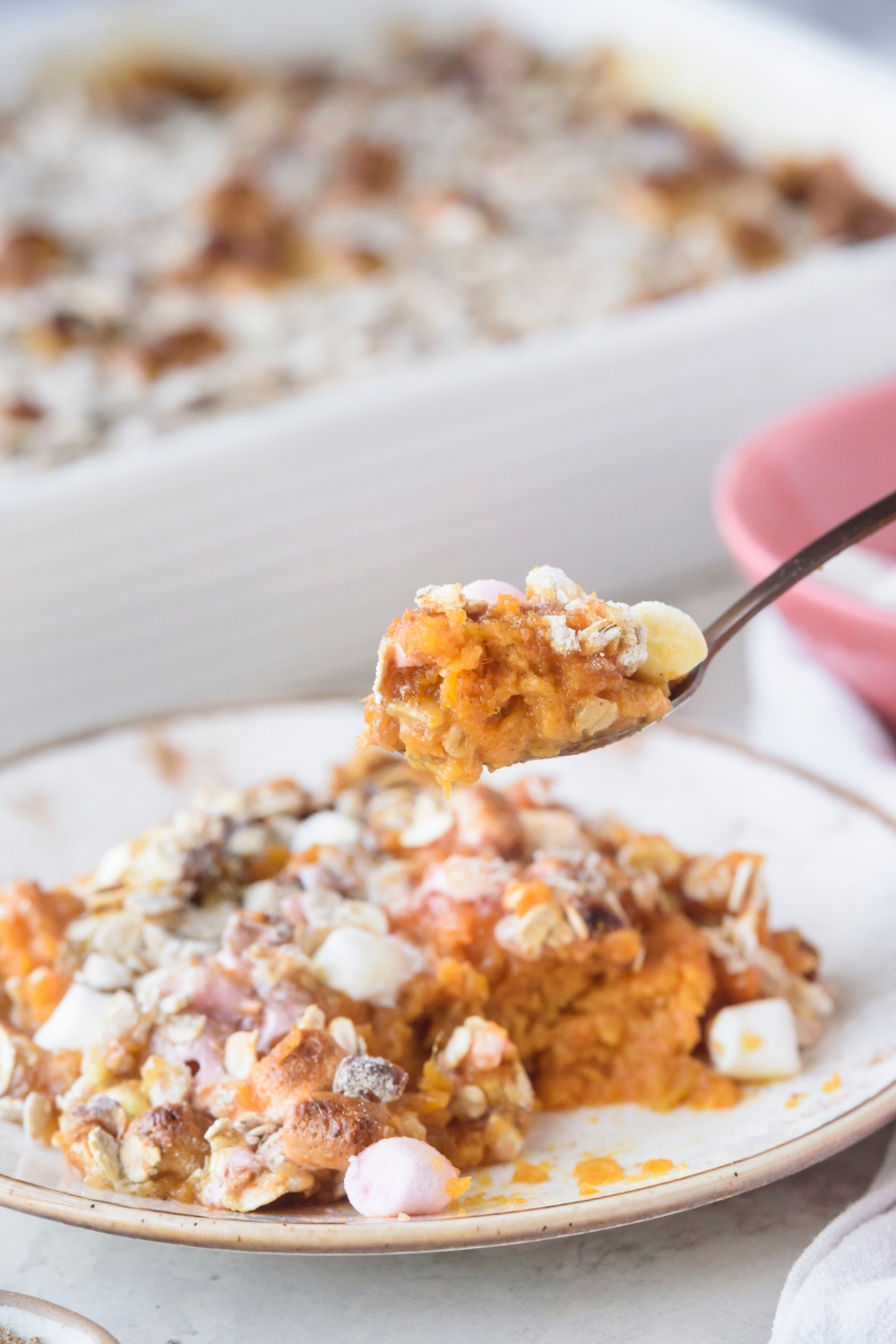 Someone lifting a spoonful of sweet potato casserole from a white plate. A full casserole dish is in the background.