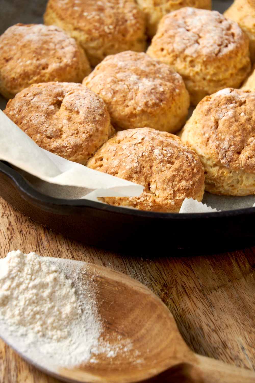 Golden brown cathead biscuits in a parchment paper lined pan with a wooden scoop filled with flour nearby.