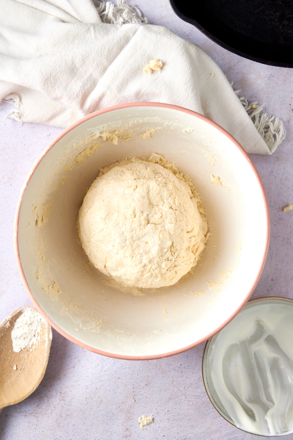 A ball of cathead biscuit dough in a white mixing bowl on a white counter with a dishtowel and wooden spoon nearby.