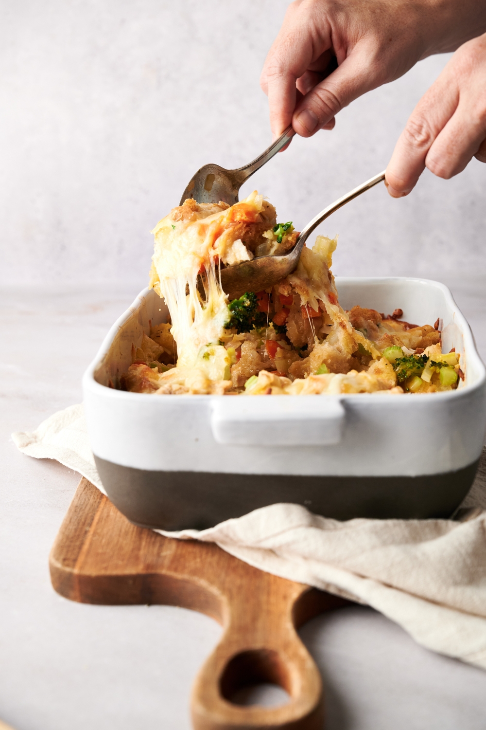 A spoon and fork dishing out a cheesy portion of chicken broccoli stuffing from a white and gray casserole dish.