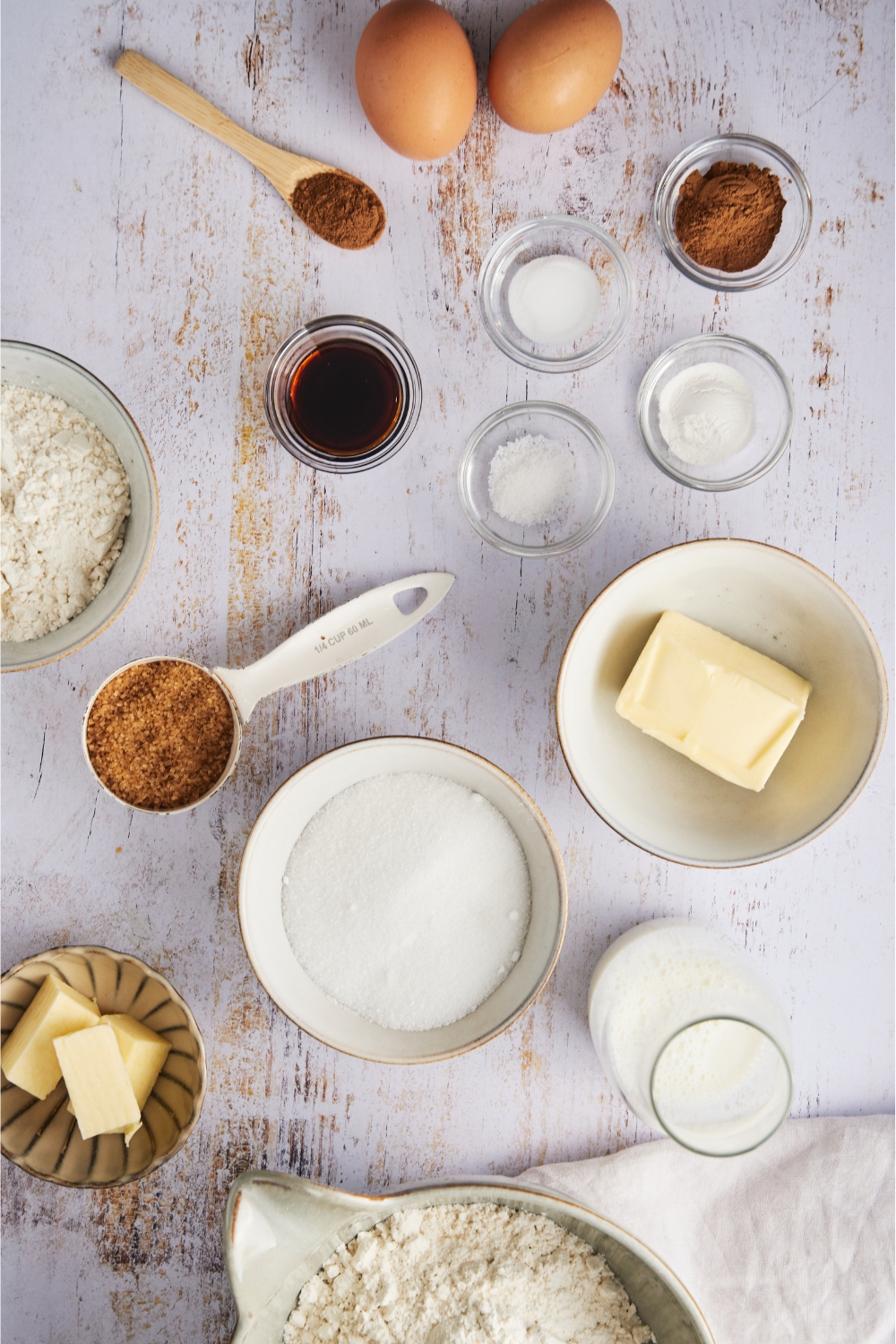 A countertop with various bowls containing the ingredients to make cinnamon streusel muffins. eggs, spices, salt, vanilla extract, brown sugar, white sugar, butter, milk, and flour.