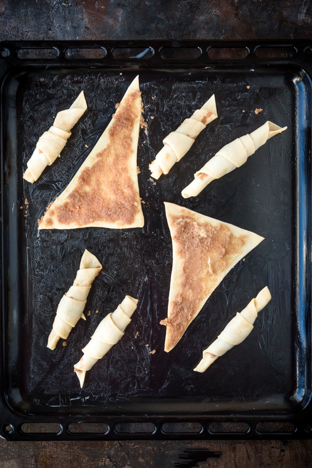 Six crescent rolls are rolled up on a black baking sheet while two triangles of dough lay flat. The triangles are spread with cinnamon and sugar filling.