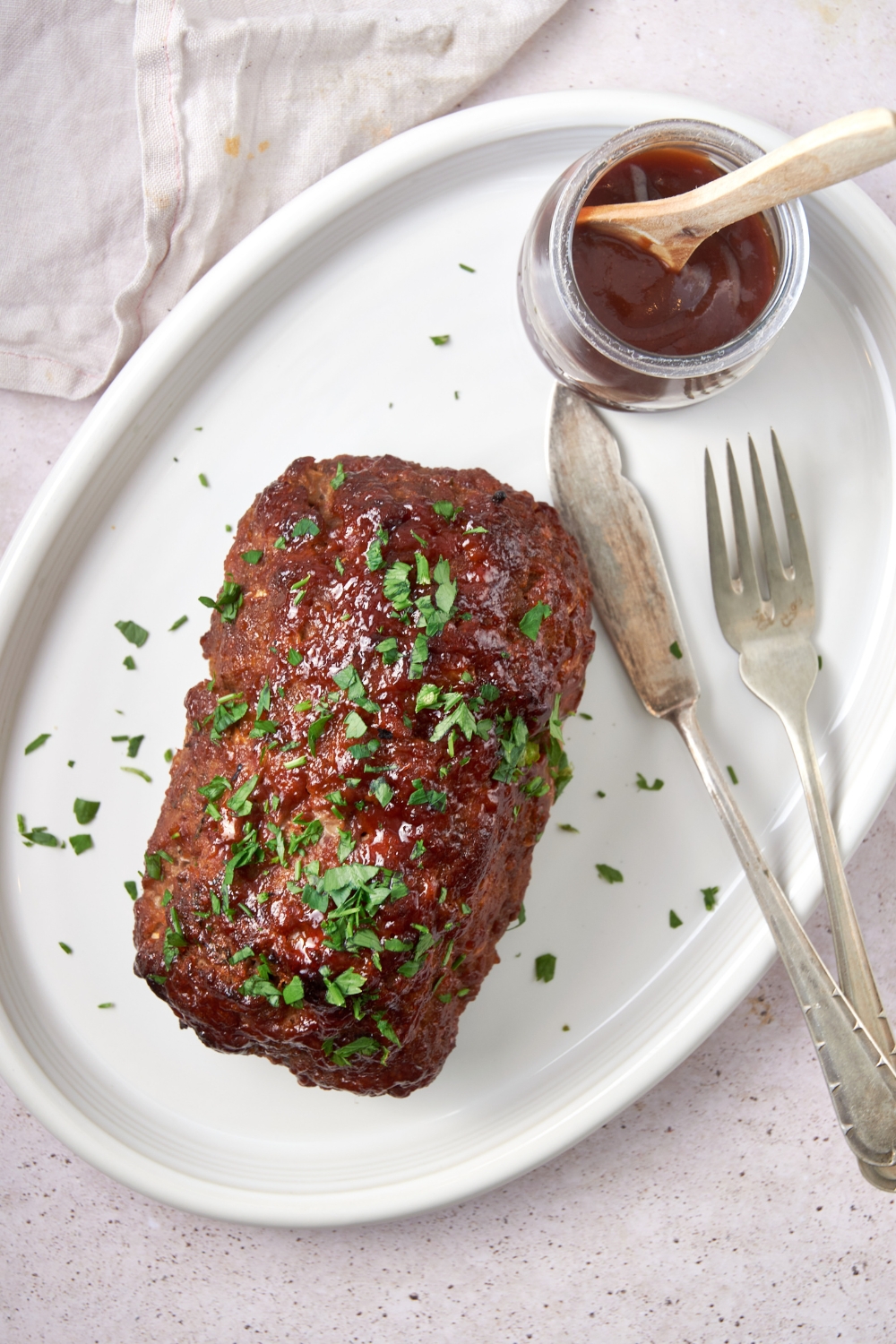 A cooked barbecue meatloaf garnished with parsley on a white serving tray. A knife and fork lay next to it with a glass jar of barbecue sauce with a spoon in it.