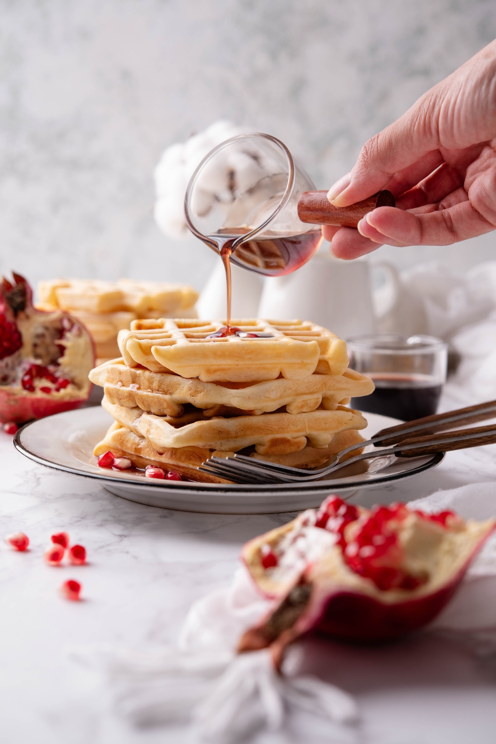 A hand pouring a jar of maple syrup over top of four waffles stacked on top of each other. There are two forks on the plate.