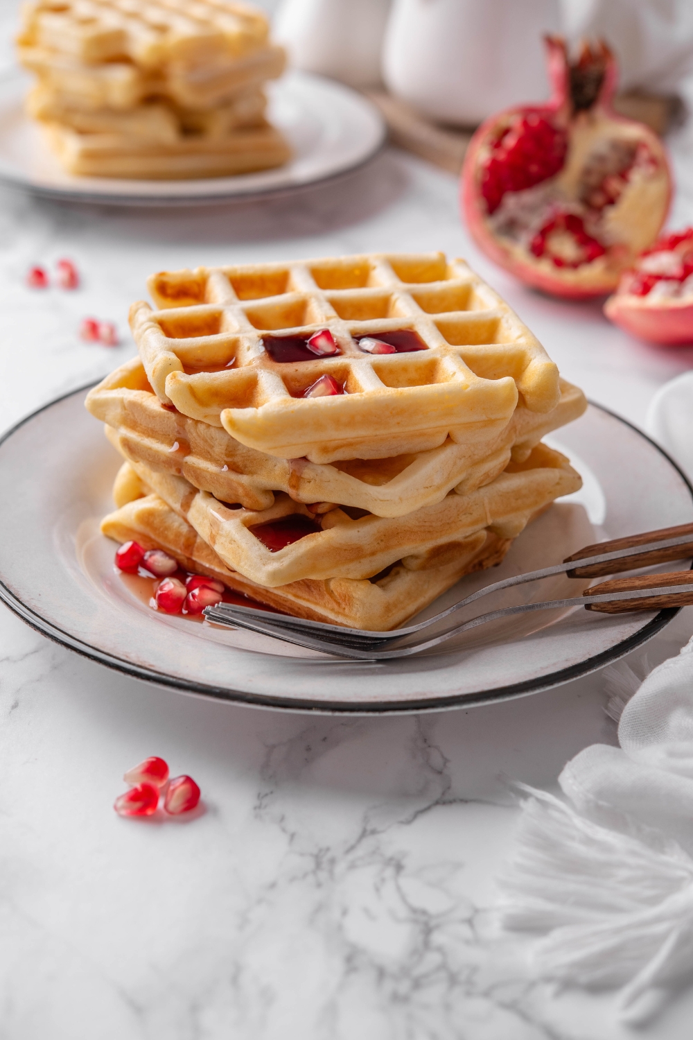 Four waffles stacked on top of each other with maple syrup dripping down the sides of the waffles and pomegranate seeds sprinkled on top and around the waffles.