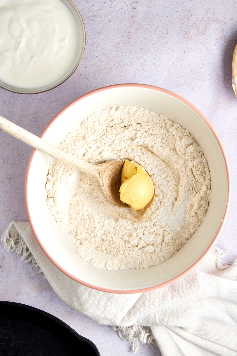 Shortening on a wooden spoon in a white mixing bowl full of flour. The bowl sits on a white counter with a bowl of buttermilk and a dish towel nearby.