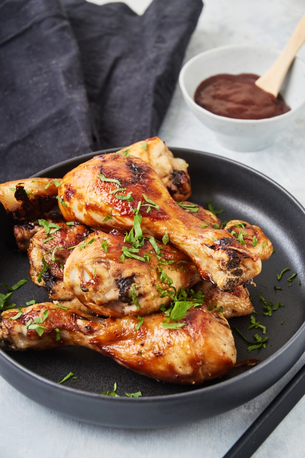 A pile of barbecue chicken drumsticks on a black plate garnished with fresh herbs and a bowl of barbecue sauce is next to it.