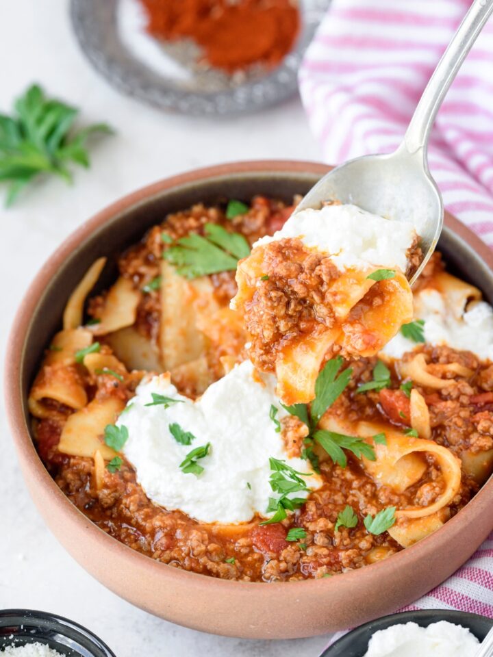 A spoonful of lasagna soup with cooked pasta, cheese, and ground beef in red sauce. The spoon is held above a bowl filled with lasagna soup.