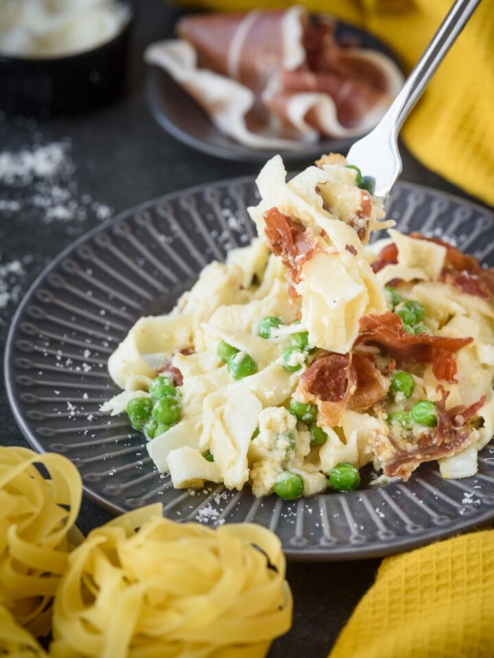 A fork holding a bite of cooked pasta with prosciutto covered in cream sauce above a plate filled with more pasta.