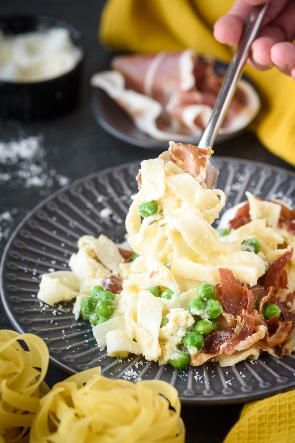 A fork scooping a bite of pasta covered in cream sauce from a plate filled with pasta, prosciutto, and peas.