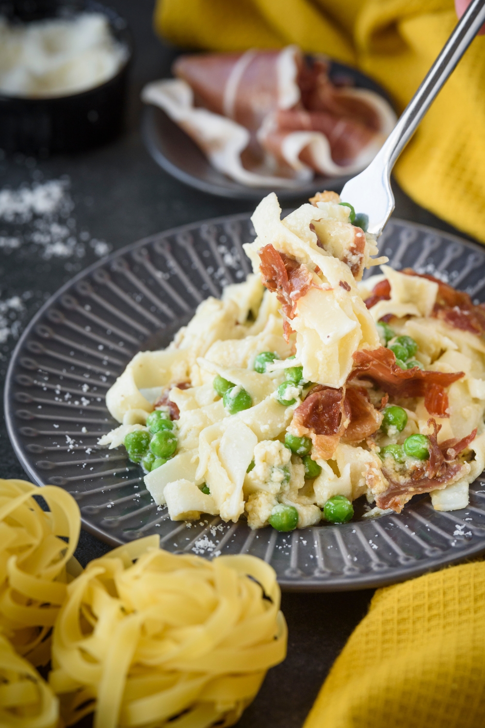 A fork holding a bite of cooked pasta with prosciutto covered in cream sauce above a plate filled with more pasta.