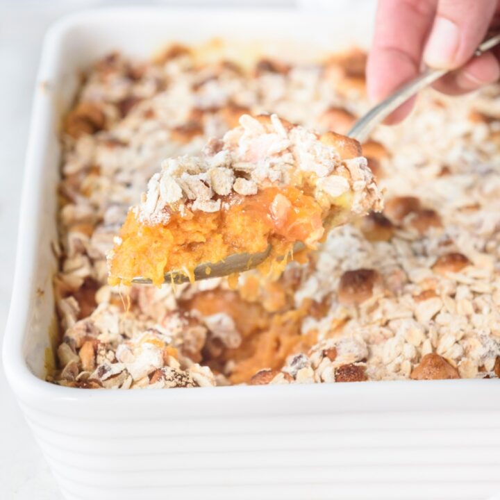 Someone is scooping sweet potato casserole out of a white casserole dish with a spoon.