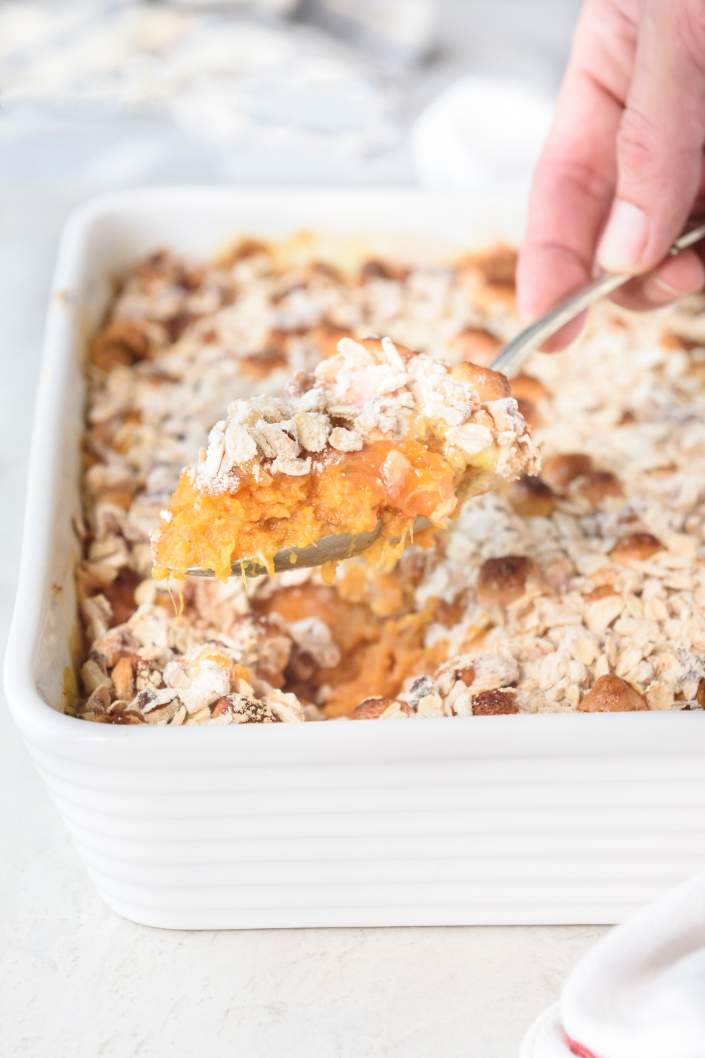 Someone is scooping sweet potato casserole out of a white casserole dish with a spoon.