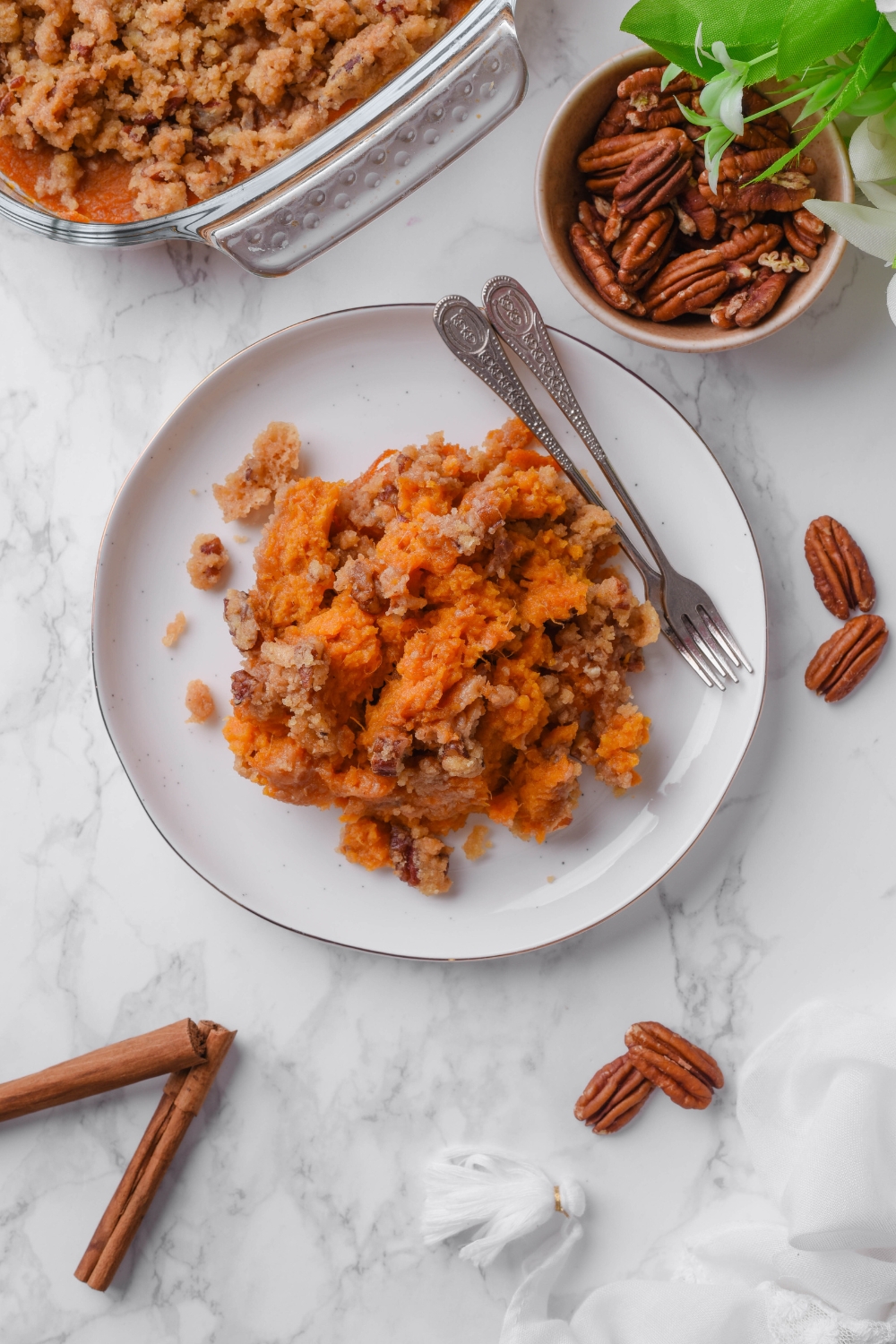 Overhead view of a plate filled with sweet potato casserole mixed with pecans and brown sugar. There are two forks on the plate and a bowl of pecans next to it along with the rest of the casserole.