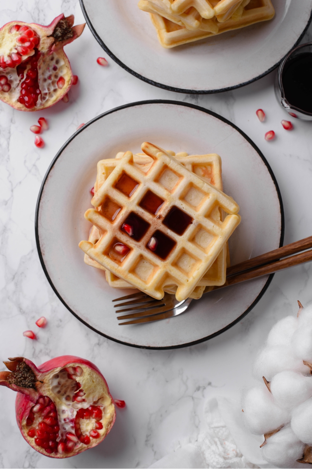Overhead view of a plate of waffles stacked high covered in maple syrup with pomegranate seeds sprinkled on top and around the counter.