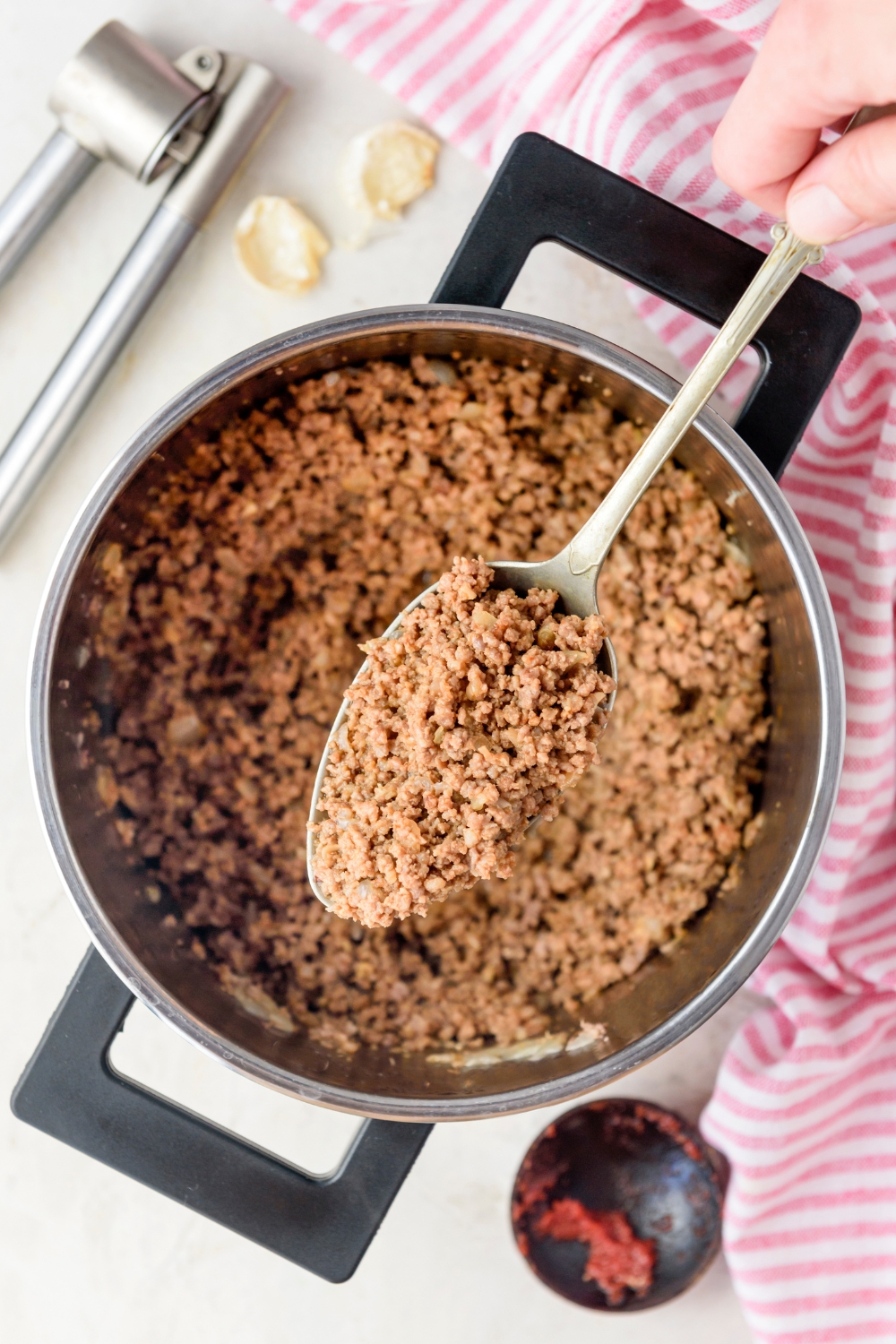 A spoonful of seasoned ground beef mixed with onions being held above a pot filled with ground beef.