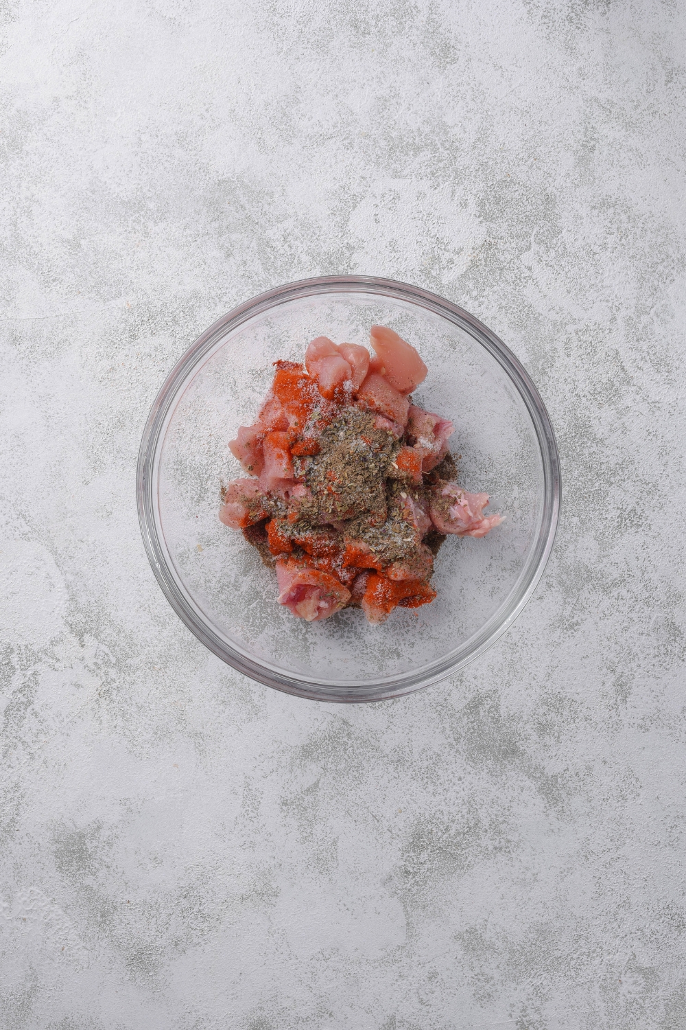 A clear bowl filled with raw diced chicken mixed with spices.