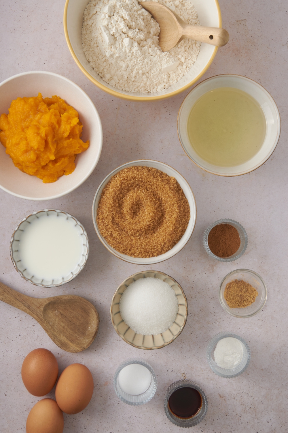 Flour, pumpkin puree, oil, brown sugar, granulated sugar, spices, and milk in separate bowls. Three eggs and a wooden spoon are also there on a white counter.