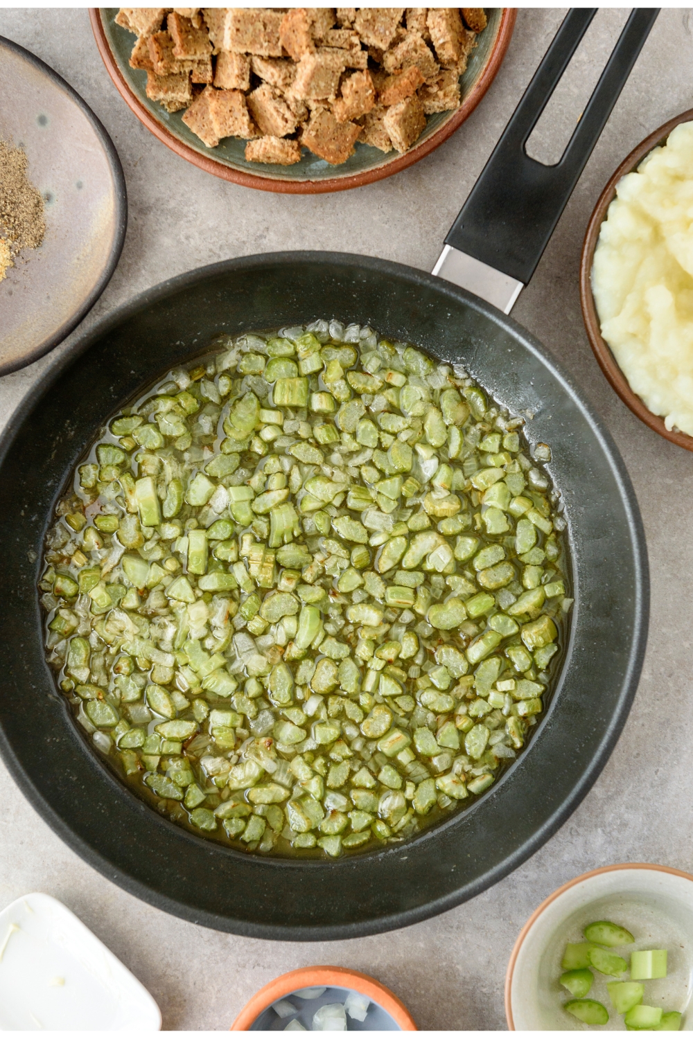 Diced celery in a black frying pan. Bowls of diced bread and potatoes are nearby.