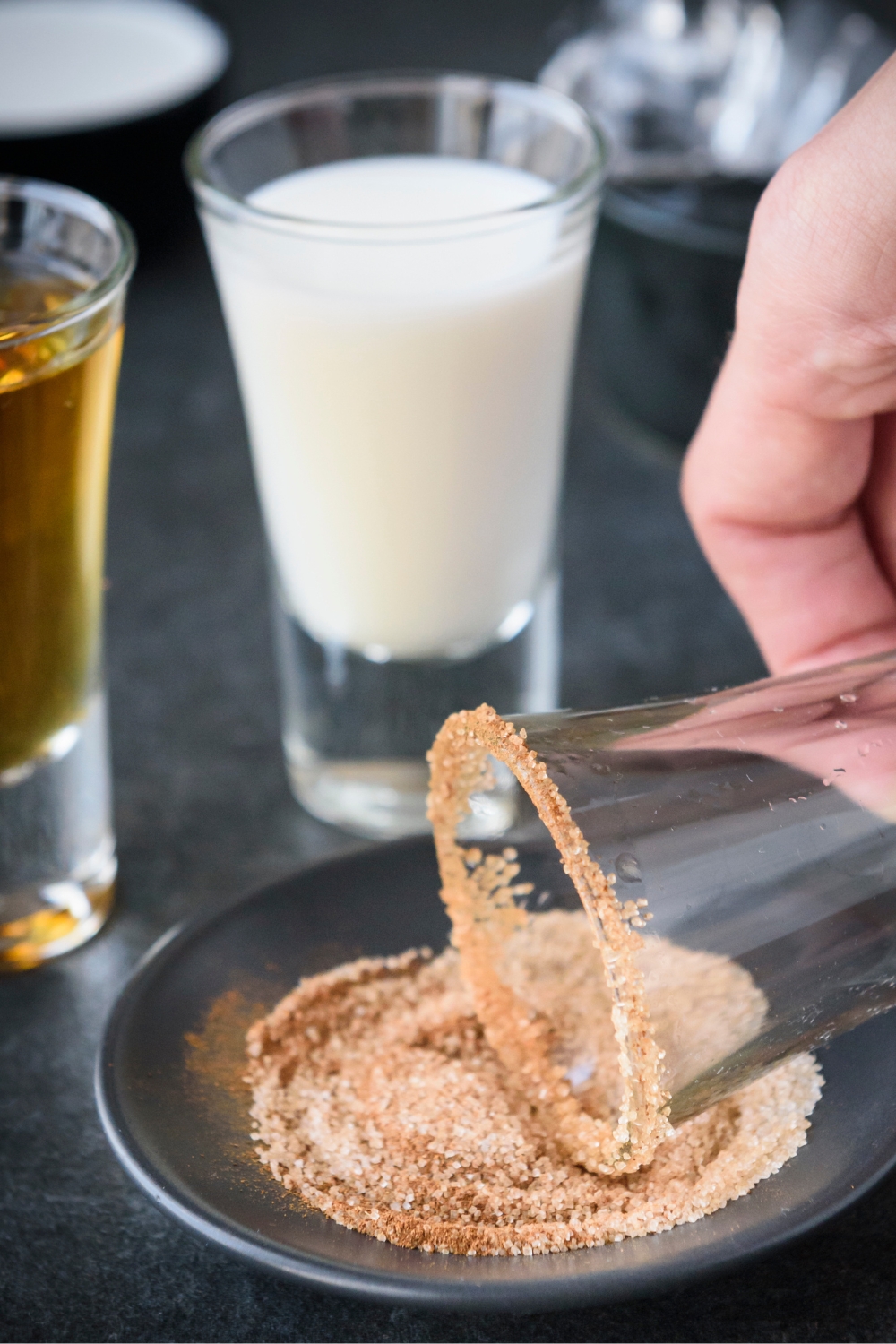 A shot glass being rimmed with cinnamon and sugar mixture. A shot glass full of Rumchata and a shot glass full of fireball stand nearby.