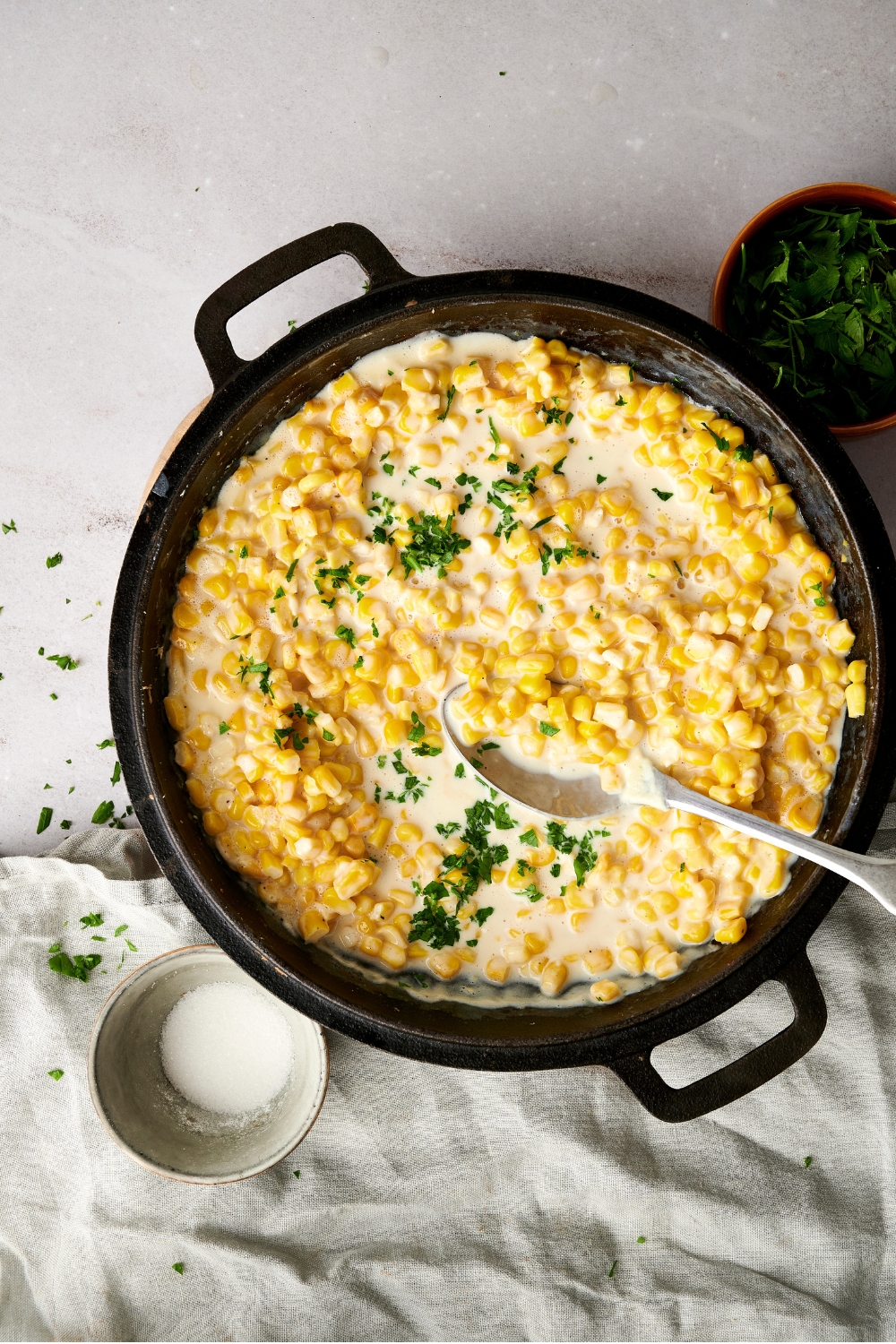 A large black pot full of creamed corn and fresh herbs with a spoon on a gray counter.