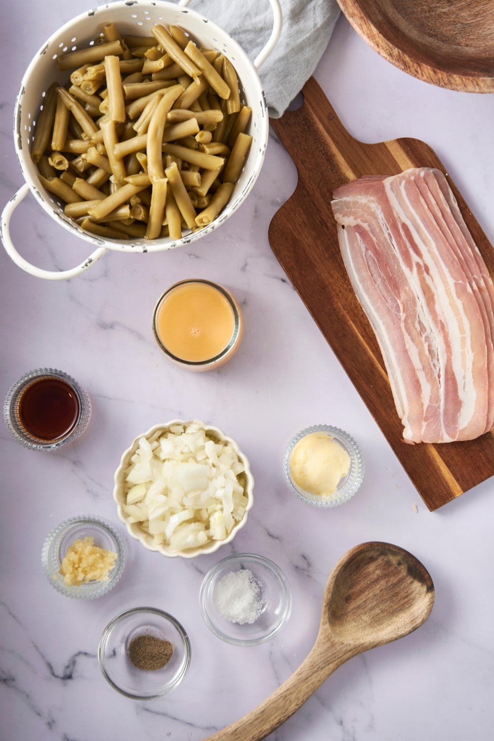 A colander of green beans, bacon, onions, seasonings, chicken broth, garlic, and sugar are in separate bowls on a white counter. A wooden spoon lays nearby.