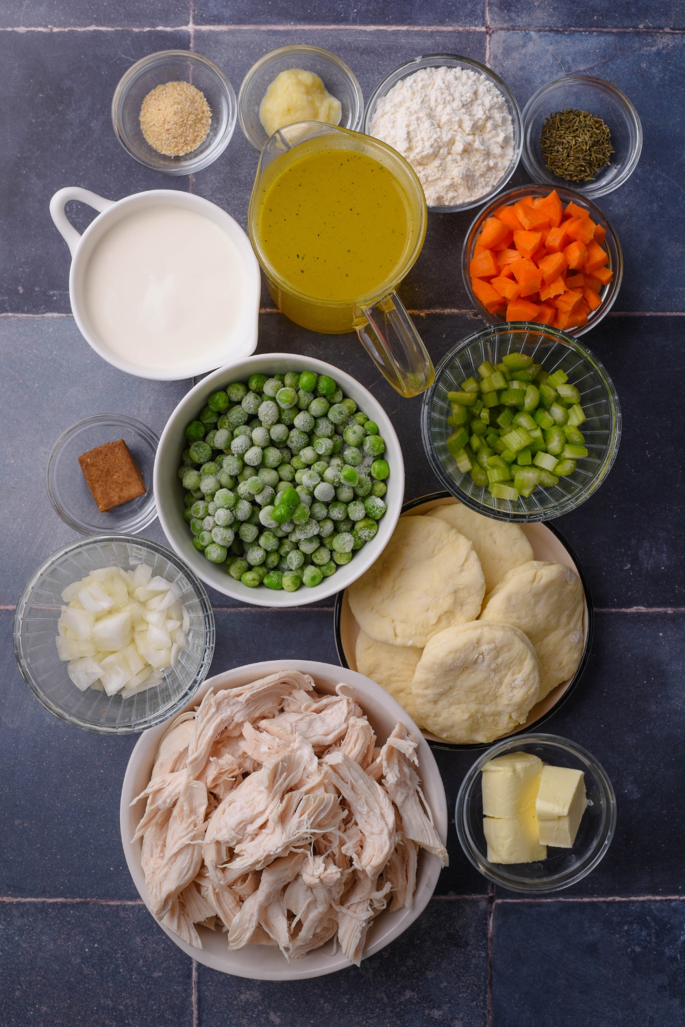 Shredded chicken, biscuits, onion, frozen peas, chopped celery, carrots, chicken broth, heavy cream, seasonings, and butter in separate bowls on a black counter top.