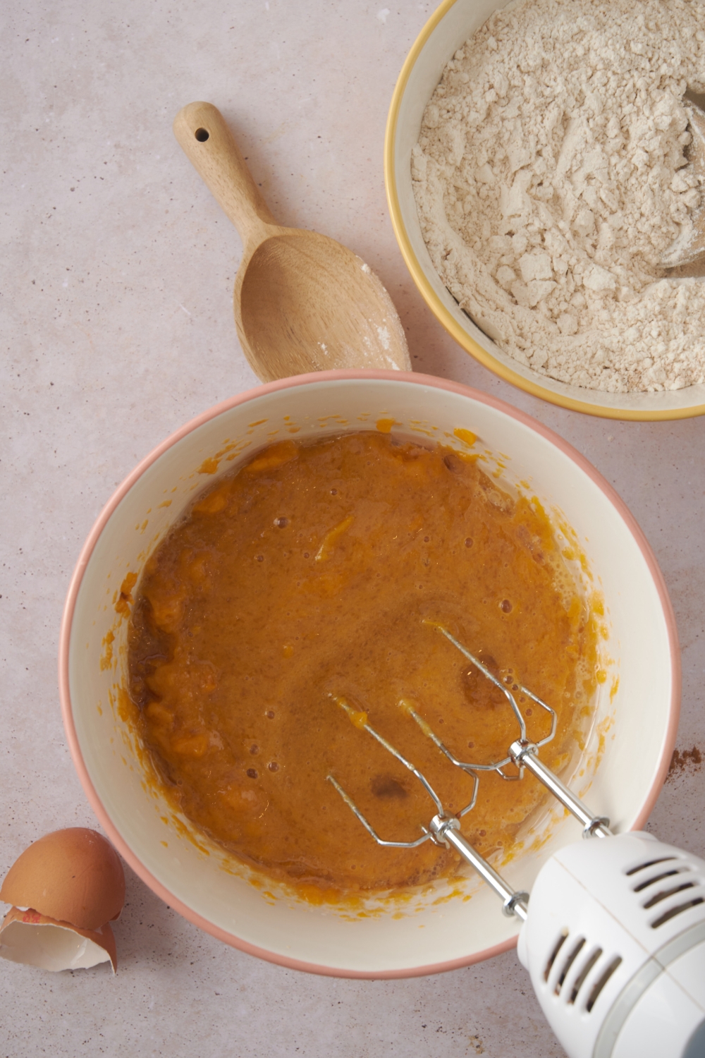 An electric mixer is combining the wet ingredients in a white mixing bowl. A bowl of flour and a wooden spoon are nearby.