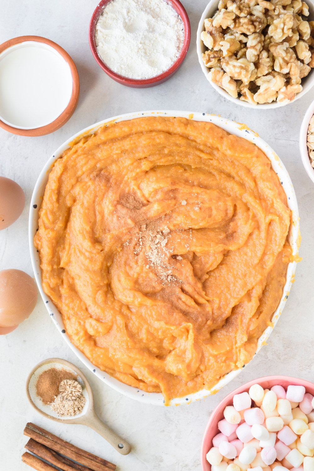 A large bowl of mashed sweet potatoes with spices on top on a white counter. Bowls of walnuts, flour, cream, 2 eggs, spices, and mini marshmallows surround the bowl.