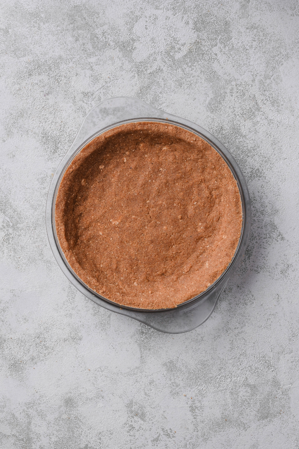 A clear pie pan with a graham cracker crust in it.