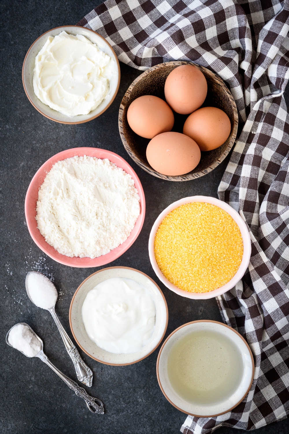 Eggs, cream cheese, flour, sour cream, cornmeal, and seasonings in separate bowls on a black counter top.