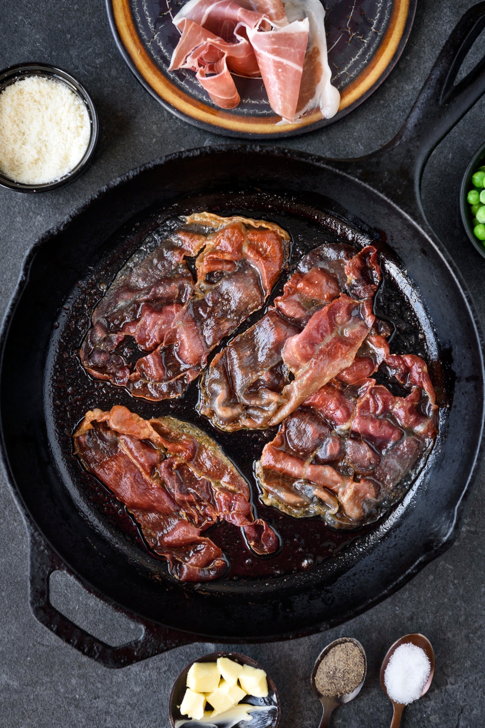 A cast iron skillet with four prosciutto slices frying in it.