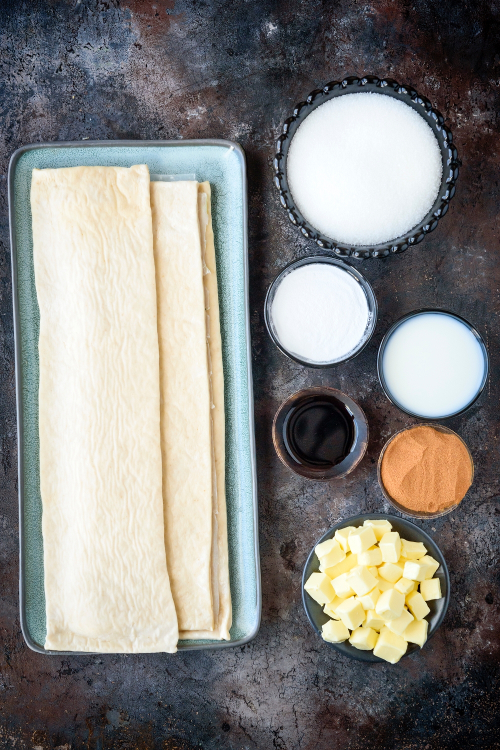 Crescent dough, milk, powdered sugar, white sugar, cinnamon, butter, and vanilla in separate bowls on a black counter top.