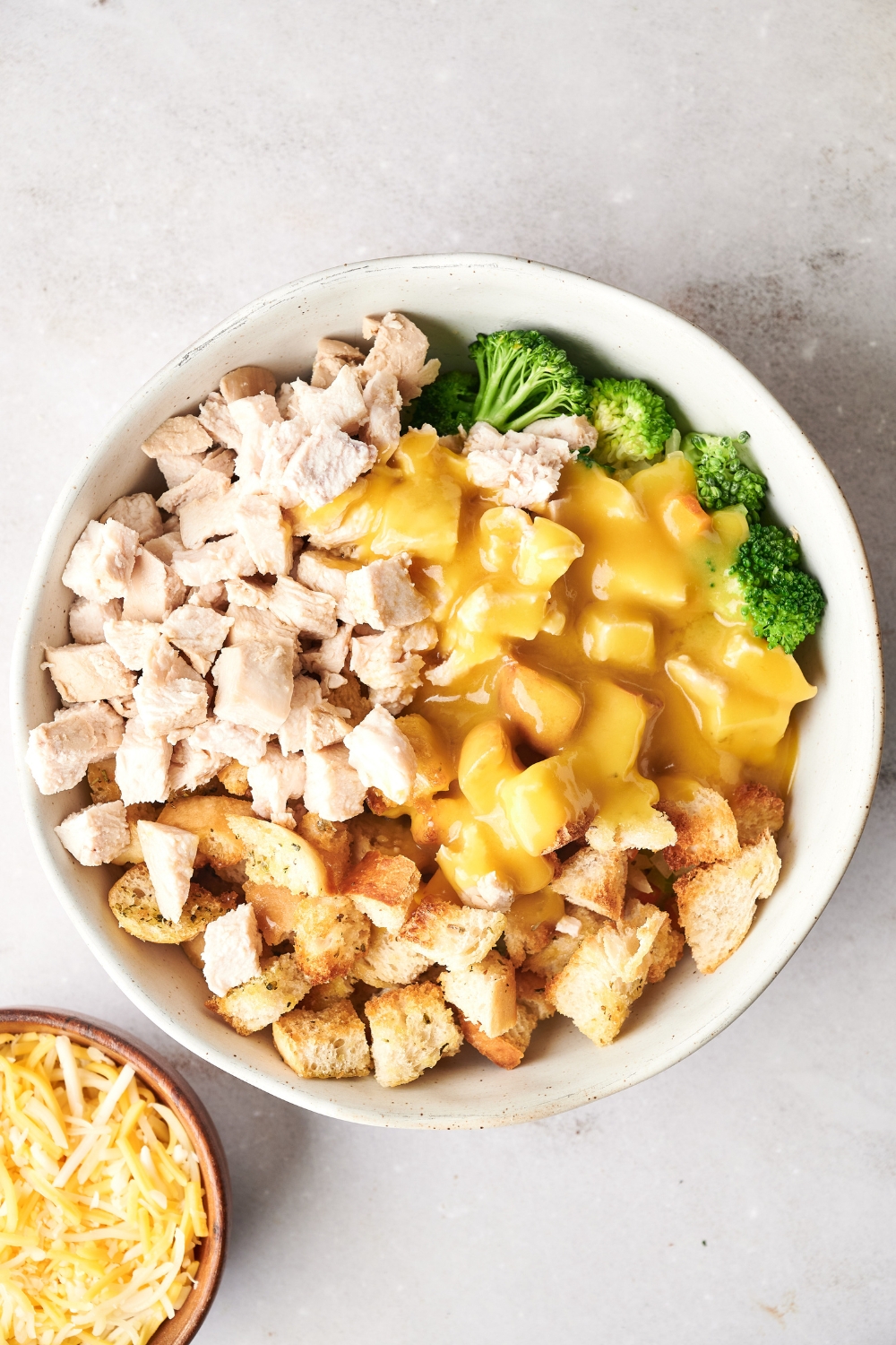 Diced chicken, stuffing, chicken soup, and broccoli are in a white mixing bowl. A bowl of shredded cheese is nearby.