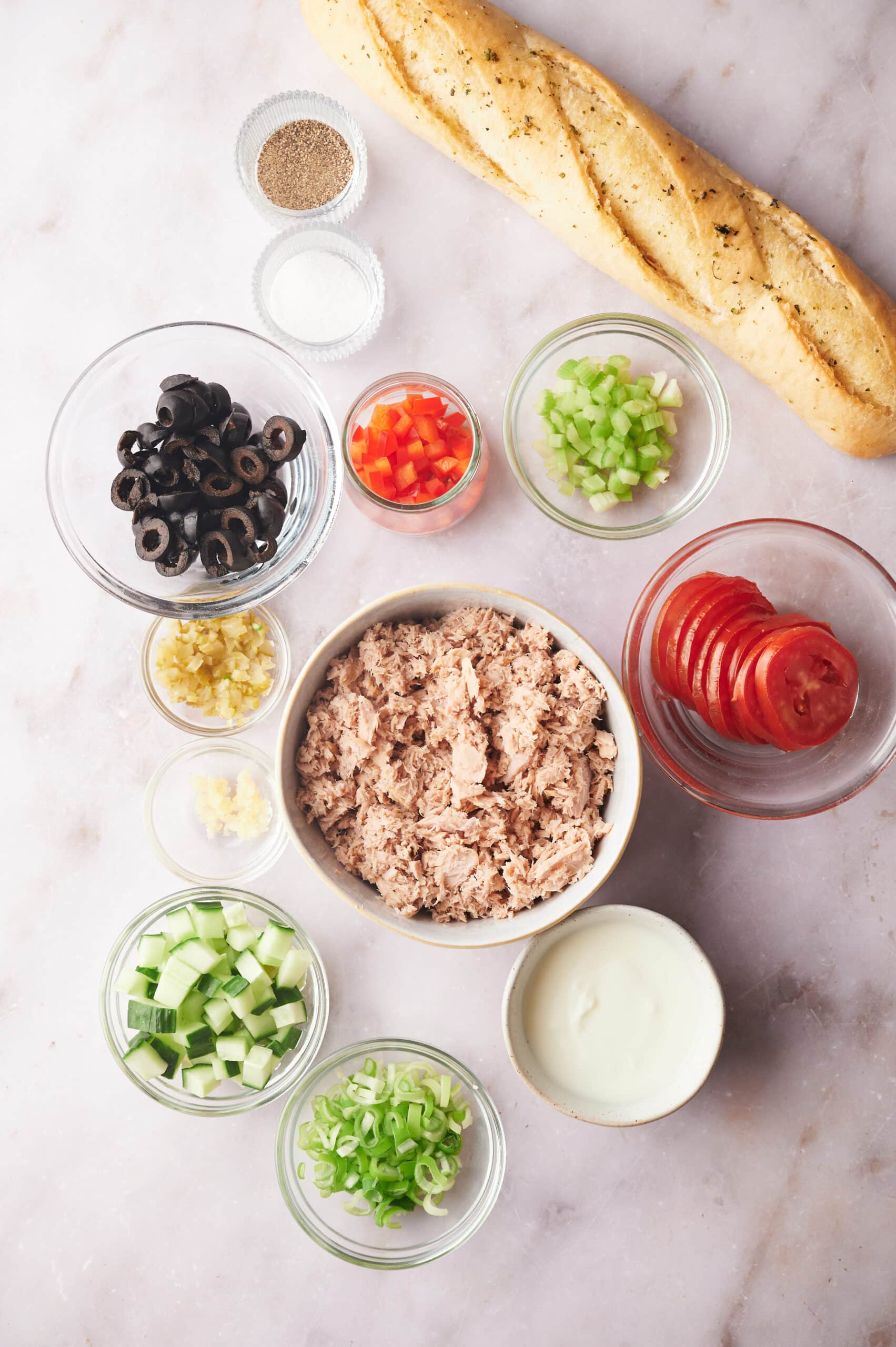 a baguette, celery, red bell pepper, sliced tomatoes, tuna, mayonnaise, black olives, and cucumber in separate bowls on a gray counter.