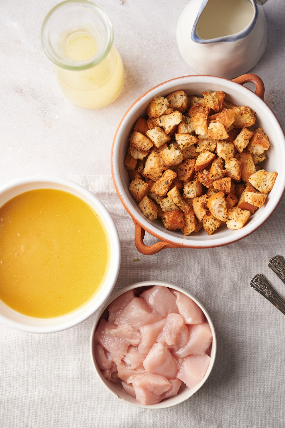 An assortment of ingredients including bowls of raw chicken, condensed soup, stuffing mix, a pitcher of cream, and a jar of broth.