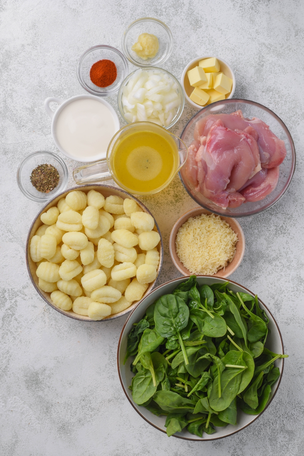 An assortment of ingredients including bowls of raw chicken, gnocchi, spinach, shredded cheese, diced onion, cream, butter cubes, and a pitcher of broth.