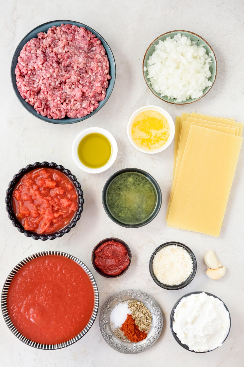 An assortment of ingredients including bowls of tomato sauce, crushed tomatoes, spices, raw ground beef, cheese, diced onion, oil, melted butter, broth, and a stack of lasagna pasta sheets.